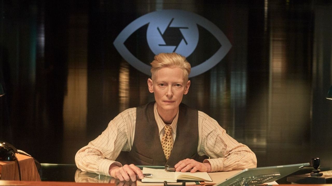 Tilda Swinton sitting at a desk by a large eye